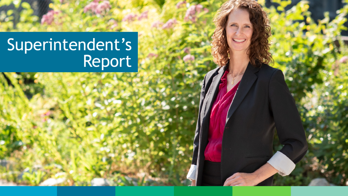 Smiling caucasian woman with brown curly mid-length hair in black blazer and magenta blouse standing in a relaxed pose in front of bright green and flowering bush in the sunshine.