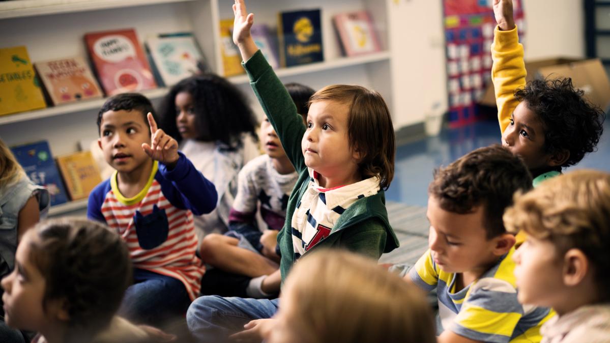 Share your ideas! Grade 1 student with hand raised among group of classmates seated on floor.