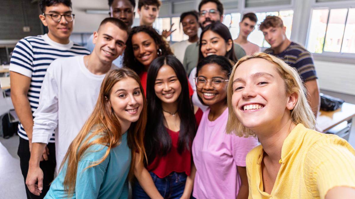 Selfie taken of smiling, diverse group of teens in classroom with teacher in background