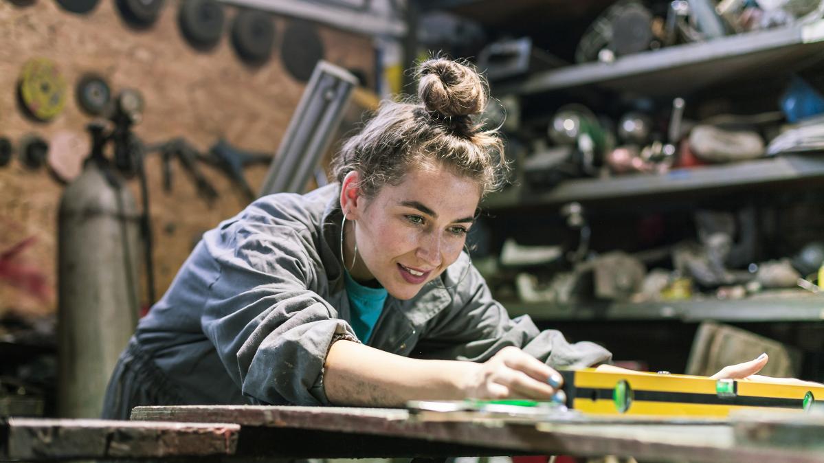 Young woman with hair in top knot wearing coveralls leans over a bench to measure something using a specialized tool