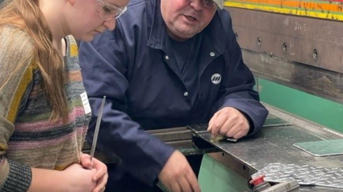 Male tradesperson trains young female trades student to operate a machine panel in an industrial setting.