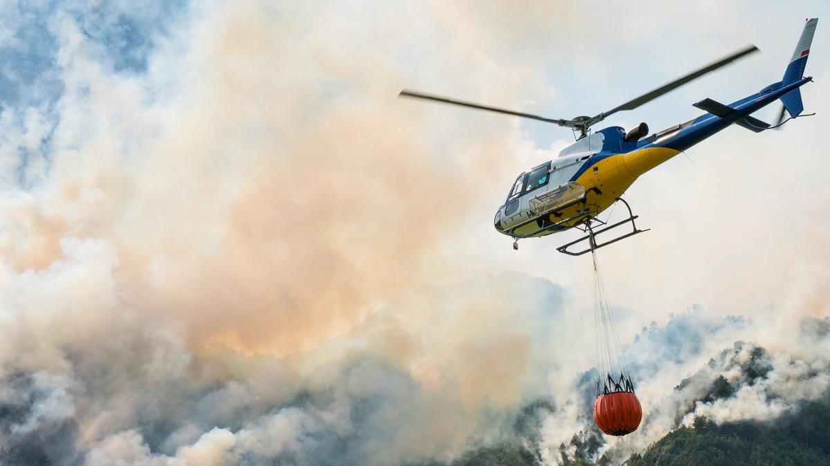 A white and yellow helicopter flying with an orange water bladder toward a large wildfire.