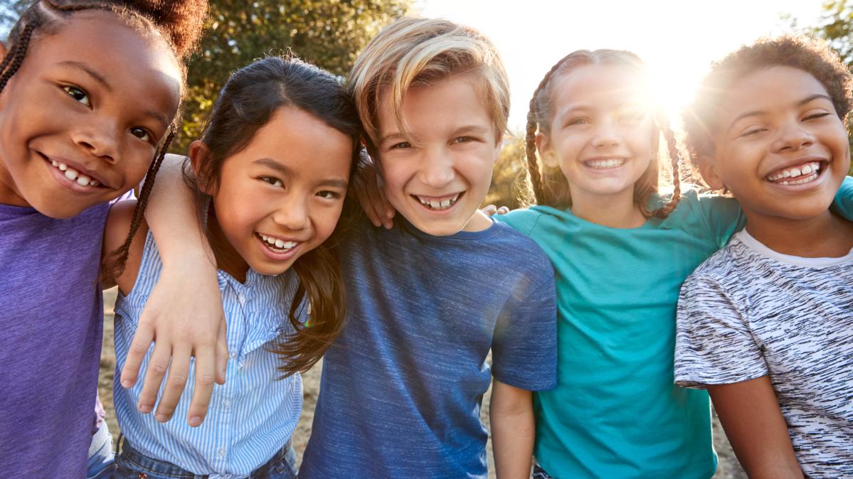 A group of diverse children arm in arm against a backdrop of sunlit trees