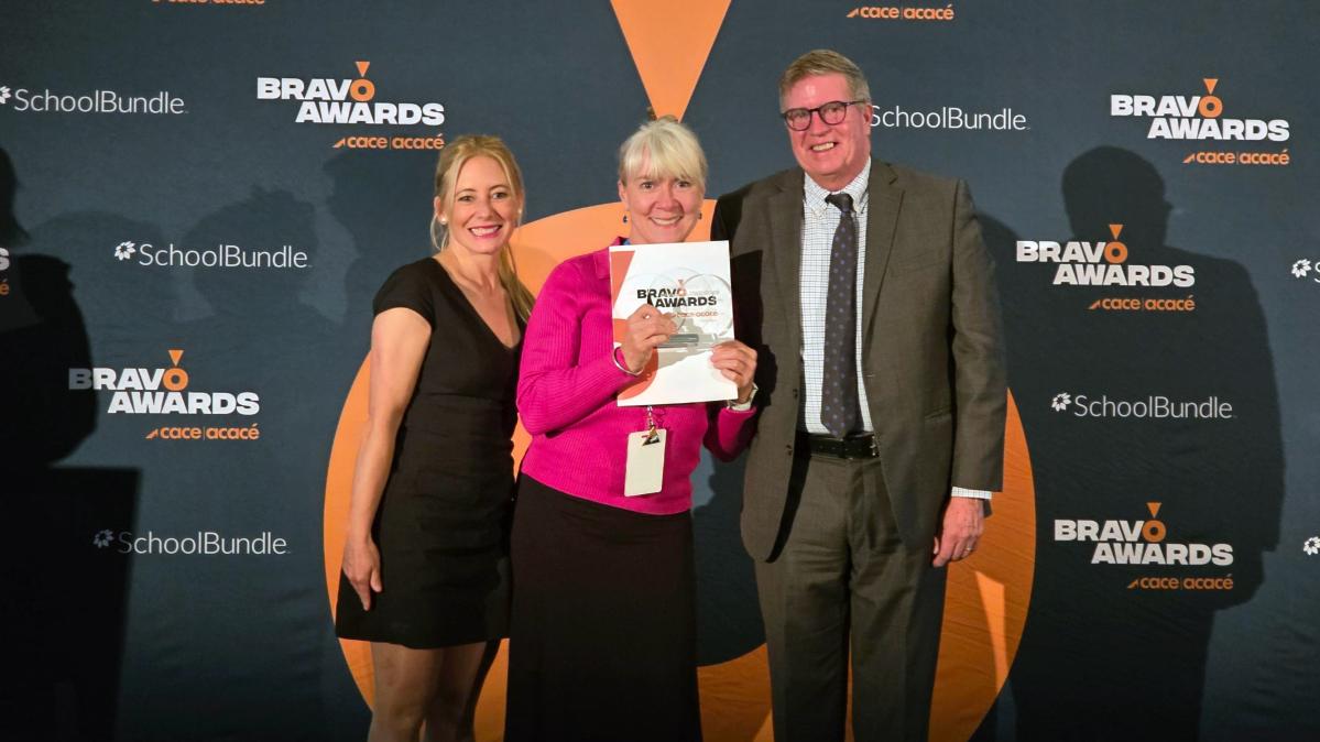 Communications & Engagement Manager Frances Maika accepts a BRAVO! award from CACE/ACACÉ President Pascale Cyr & SchoolBundle's Jacques Major Two women in dresses and a man in a suit pose in front of a grey wall with the middle woman holding up a certificate.