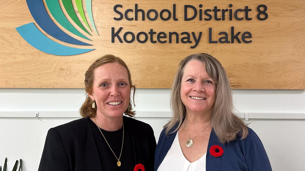 A woman with red hair in a black suit stands beside a woman with blonde, shoulder-length hair in a blue sweater and white shirt, in front of a wooden sign indoors.