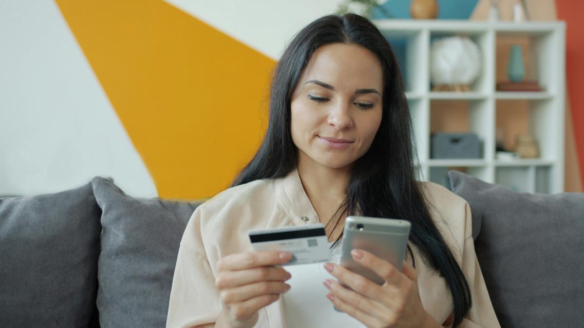 A woman with long, dark hair and a tan blouse sits on a grey couch holding a credit card and a cell phone. She is looking at the cell phone as if making an online payment.