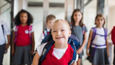 Young, grinning elementary school student in red sweater vest, white t-shirt and navy blue backpack runs toward the viewer with his classmates behind him in a bright, daylit school hallway