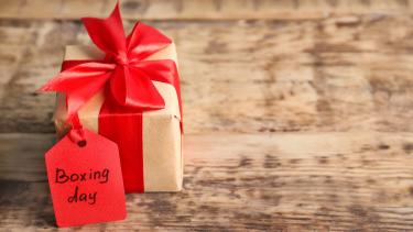 Boxing Day Photo of smallish gift wrapped in brown paper with red ribbon and red label with boxing day written on it in black pen, with the gift sitting on a weathered, wooden plank background that could be a floor or a table top.