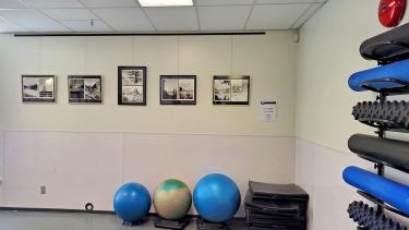 A fitness centre with rolled up mats along a wall on the right along with blue and tan fitness balls of various sizes on the back wall above which are hanging ink paintings in black frames against a white wall.