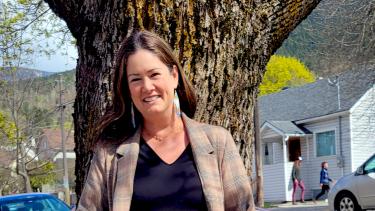 Woman with brown hair in tweed blazer, black shirt and blue jeans standing in front of the trunk of a maple tree on a sunny day.