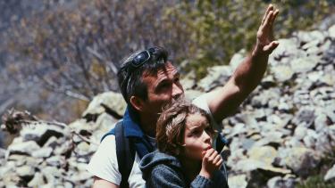 Man gesturing toward rock face with young child sitting in front of him as if explaining something to the child.