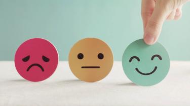 Mental Health A red frowning face, a yellow neutral face and a green happy face cardboard cutouts in a row with a hand picking up the green one.