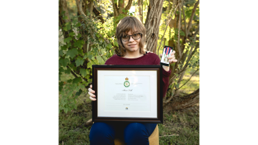 Alexis Folk, 2024 King Charles lll Award Recipient A young woman with wavy blonde hair wearing a burgundy sweater seated on a chair in a forest holding a medal and a framed certificate.