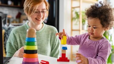 Young woman looks on, smiling, as a toddler builds a tower of coloured blocks.