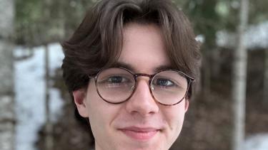 A teenaged boy with brown hair wearing glasses and a green jacket stands in front of some trees.
