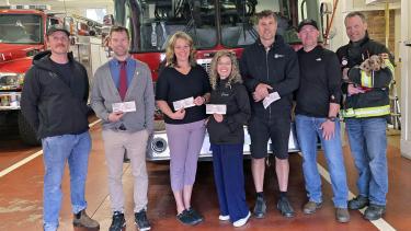 A group of people stand in front of a fire truck inside a fire department, holding cheques in front of themselves.