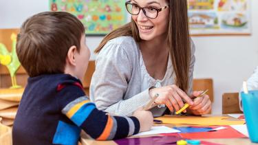 A young teacher with glasses and brown hair leans against the desk of a grade 1 boy as he looks at her and smiles widely.