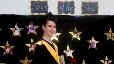 A high school graduate in cap and gown standing in front of a black backdrop decorated with gold stars.