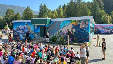 A group of elementary school students sit on the ground in front of a colourful school while a principal speaks to them.