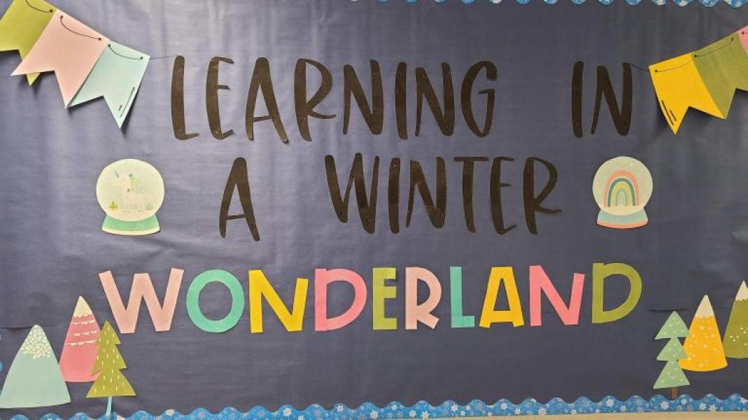 Wall-mounted homemade banner with dark grey background and multi-coloured lettering in foreground that reads, "Learning in a winter wonderland." Cutouts of prayer flags at top corners, snowglobes in middle and trees at bottom corners.