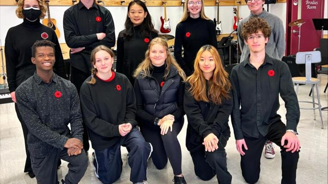 Group of students kneeling and standing in formation with black outfits and red poppies on their lapels in school band room. 