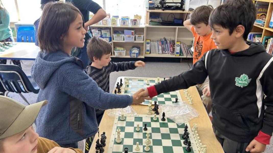 Two children shaking hands across a chessboard after a game while two other children continue to play in the background. In an elementary school setting with bookcases against a back wall and light streaming in from a window in the background.