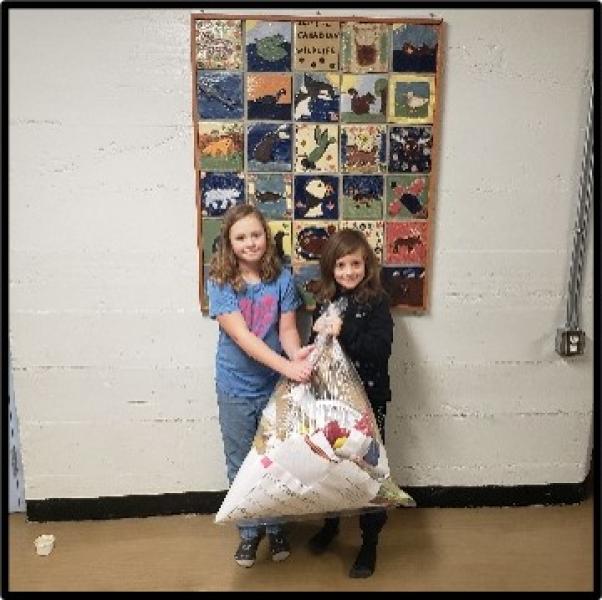Two elementary aged girls standing in a hallway in front of a wall collage holding a bag of recycled paper between them.