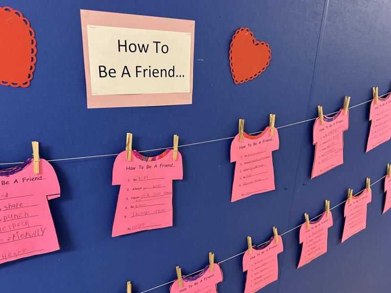 Cutouts of pink t-shirts with messages written on each describing, "How to be a friend" and each cutout attached to a line by a wooden clothespin and hung on a blue wall with hearts also taped to the wall. 