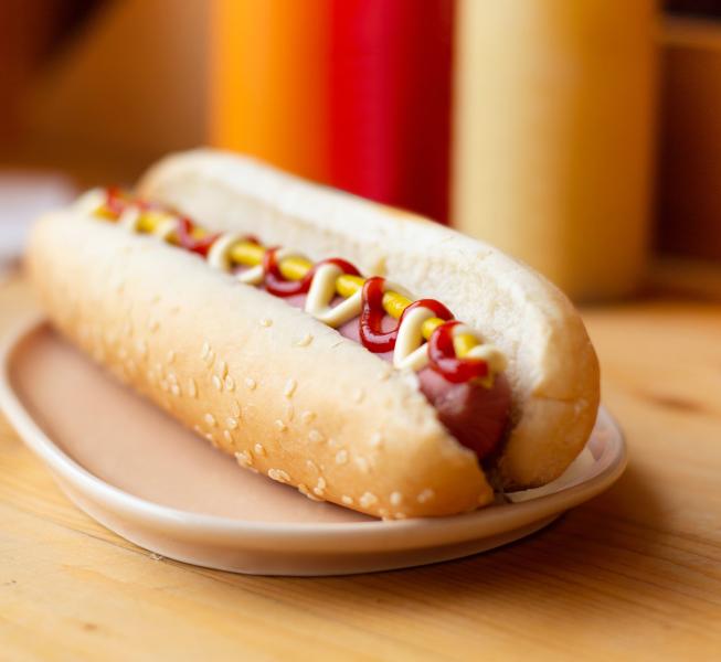 Hot dog with mustard, mayo and ketchup on a sesame seed hot dog bun on a beige plate on a wooden countertop with condiment bottles in background