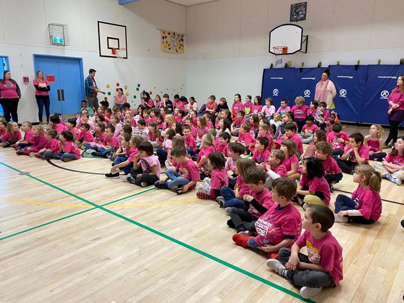 An elementary school class seated on a gymnasium floor, all wearing pink shirts.