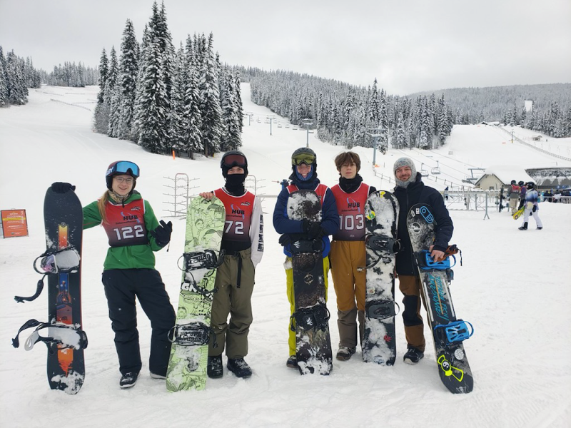 Four smiling teenaged students and their teacher/coach in winter clothing standing in front of chairlift at ski resort leaning on their snowboards.