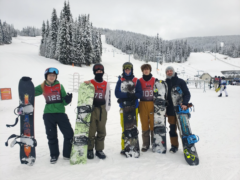 Four smiling teenaged students and their teacher/coach in winter clothing standing in front of  chairlift at ski resort leaning on their snowboards.