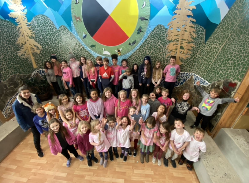 Birdseye view of elementary school class in front of a forest mural at the centre of which is a painting of a coloured circle depicting the First Nations' four directions.
