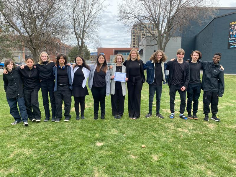 Group of students in dark winter jackets stand with arms linked on green lawn in front of a school.