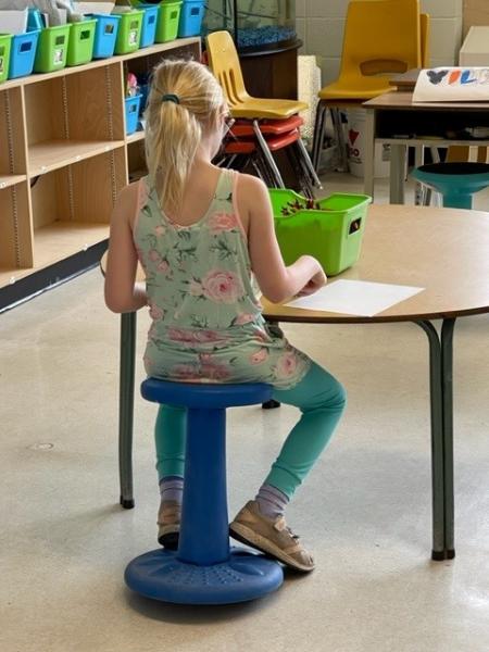 Elementary school student with blonde ponytail, green tights and flowered green tank top sits at a round table in front of a piece of paper and a green tote in her classroom. 