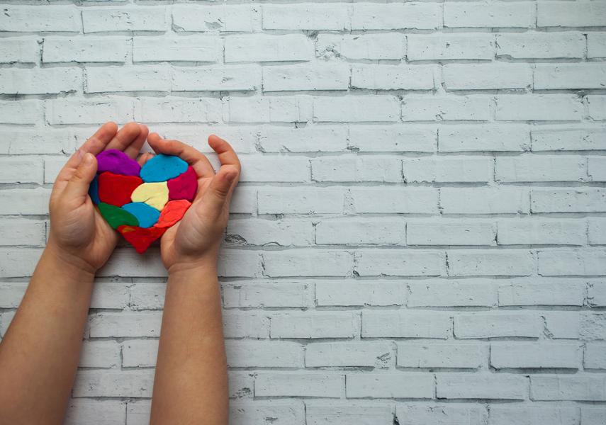 Brick wall painted light grey in background with set of child's hands holding multi-coloured plasticine molded heart on left side of photograph. 