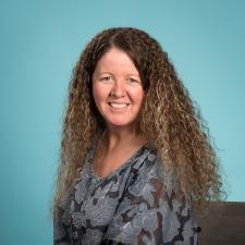 Woman with long wavy brown hair in grey shirt against teal background.