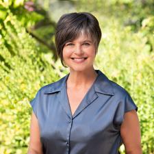 Smiling woman with asymmetrically cut, short brown hair in short-sleeved collared grey silk shirt in front of green bushes with sunlight in background