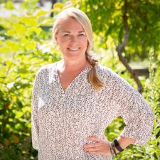 Smiling woman with blonde hair in loose braid over her should wearing finely patterned white blouse standing with one hand on hip in front of green bush and tree with sun beaming behind her.