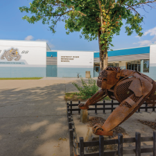 A flat-roofed blue and white school building against a blue sky with a leafy green tree and a rust-coloured sculpture of a bulldog in the foreground