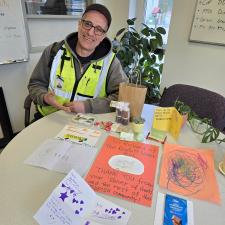 A man in a ball cap, glasses and a flourescent green vest sits at a table covered in hand-drawn cards and messages.