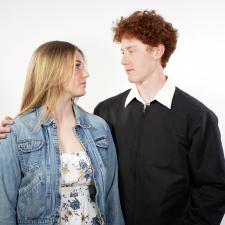 A young blonde woman in a jean jacket looks into the eyes of a young red haired man in a black zipped jacket while he has his arm around her shoulder.
